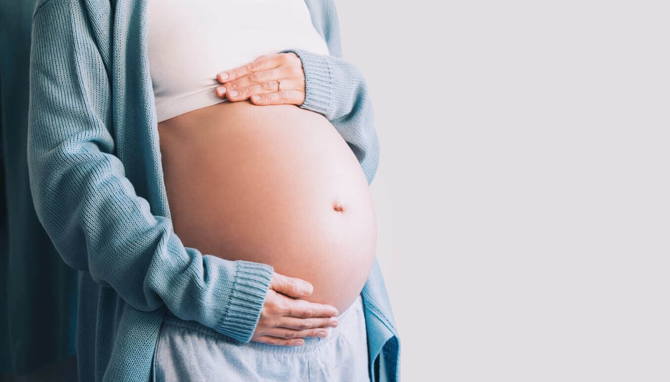 Mulher grávida com as mãos apoiadas sobre a barriga, vestindo cardigã azul e top branco